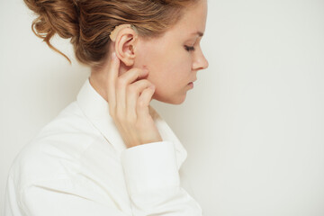 Fototapeta premium A woman touches her ear with a hearing aid in place, standing alone against a neutral background. 