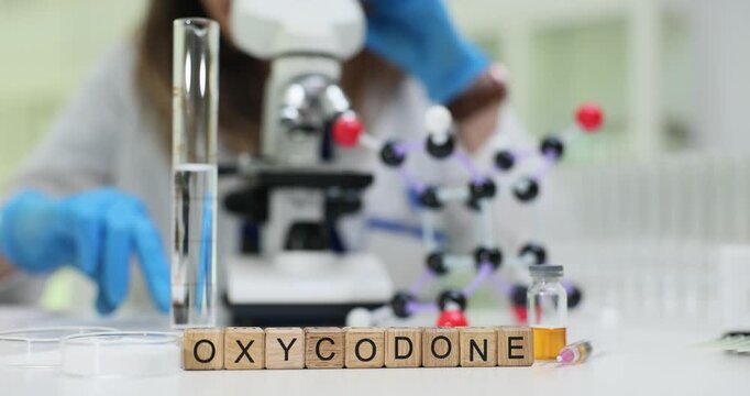 Wooden blocks spell word Oxycodone near glass tube and molecule model. Woman researcher checks opioid sample analysing strong painkiller risk