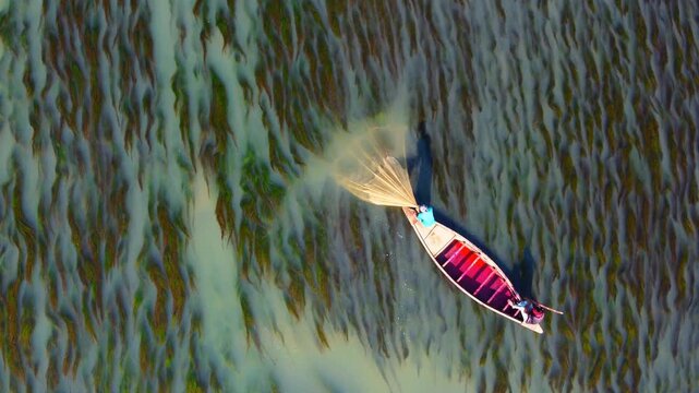 Rajshahi, Bangladesh - 15 December 2025: Aerial view of a lone fisherman in a boat casting a net, creating a circular splash against the textured riverbed.