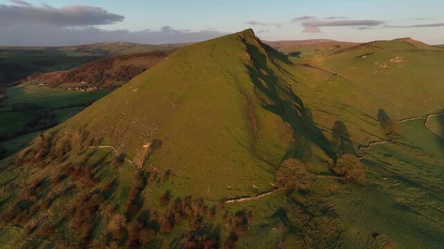 Aerial view of the vibrant green Mam Tor peak, casting long shadows across the Peak District's rolling hills, Buxton, England, United Kingdom.