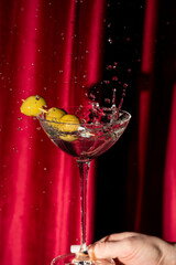 A vibrant martini cocktail with green olives is captured mid-splash against a dramatic red curtain backdrop.