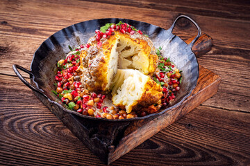 Vegetarian dish - whole oven baked roasted cauliflower with tahini sauce, chickpeas and pomegranate in rustic iron skillet pan on wooden board