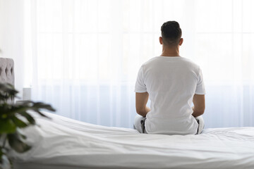 A young man in a white t-shirt sits on his bed with his back facing the viewer. He gazes out the...