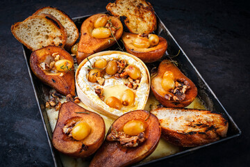 Oven baked camembert cheese with caramelized glazed pears, walnuts and grapes and baguette served as close-up on rustic metal backing sheet