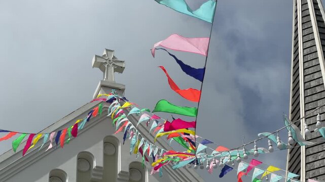 A Catholic church cross is framed by strings of bright flags and slow moving clouds, blending devotion and festive detail within a tranquil open sky background. Sacred symbol.