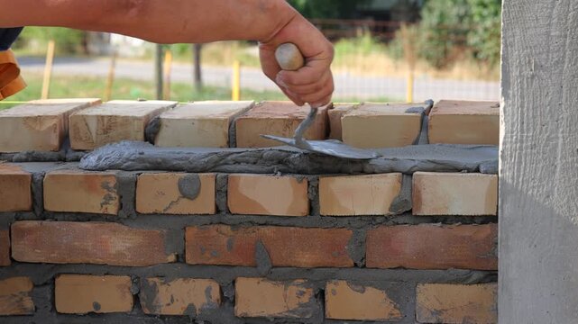Professional construction worker is laying bricks while building a wall at a construction site. Close up of bricklayer using masonry tools to apply mortar and align the bricks with precision.