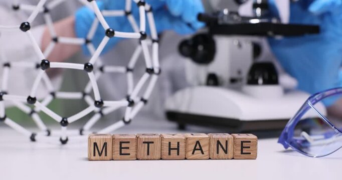 Laboratory worker uses microscope to examine sample portraying methane. Cubes arranged on table form word Methane indicating subject of analysis