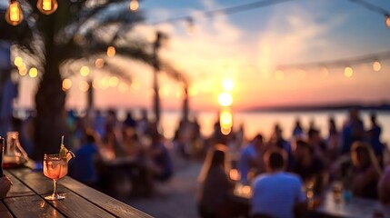 Naklejka premium Vibrant tropical cocktail with a garnish sits on a wooden bar counter while blurred people socialize at an outdoor beach club during a picturesque summer sunset