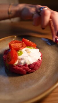 close-up of a female hand rotating a plate on a table containing a portion of meat tartare with cherry tomatoes and stracciatella