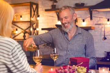 Cheerful middle-aged man pouring wine into glasses during romantic date in the kitchen. Quality time together - love language. Celebration of special event