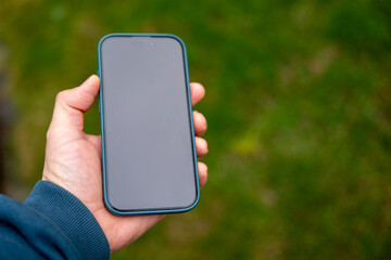 Hand holding a modern smartphone with blank screen outdoors. Mockup device with copy space, green natural background, mobile technology concept, app presentation or user interface display.
