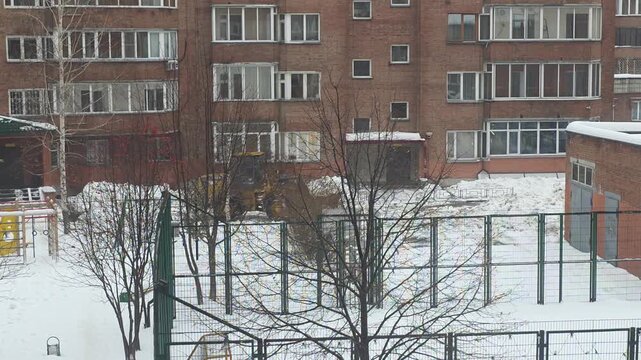 Yellow front loader clearing snow near a brick residential building and fenced playground in a snowy winter landscape