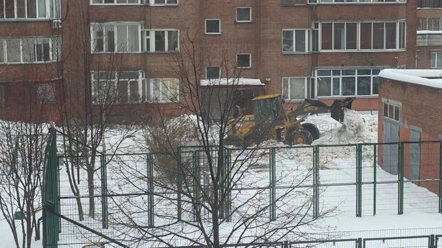Yellow front loader clearing snow near a brick residential building and fenced playground in a snowy winter landscape