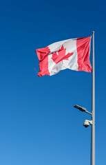 Red and white Canadian flag on a tall pole with mounted streetlight and security camera against a clear blue sky.