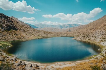 A bright blue lake rests peacefully within rugged brown mountains under a clear sky with white clouds.