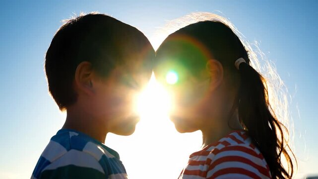 Silhouette of two children, a boy and a girl, touching foreheads with eyes closed against a bright sunny sky, conveying love and connection