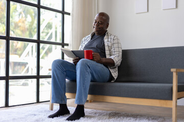 Senior man enjoying his retirement, sitting comfortably on sofa, reading interesting book and drinking coffee in cozy living room next to window.