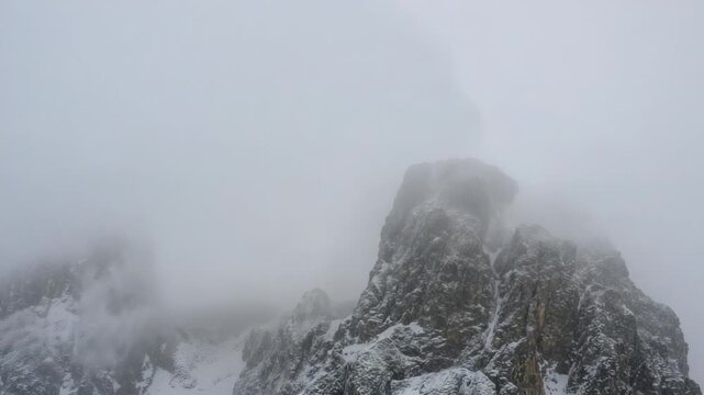 Drifting low cloud enveloping rugged snow-dusted peak at alpine ridge, revealing and veiling summit
