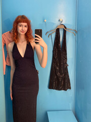Young female caucasian trying black dress in bright blue changing room mirror selfie