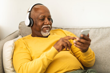 A senior man wearing headphones smiles as he listens to music on his smartphone while sitting comfortably on a sofa. The cozy living room setting adds to the relaxed atmosphere.