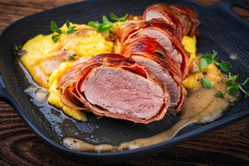 Pork fillet roast wrapped in prosciutto ham bacon with polenta and fresh herbs in serving cast iron skillet