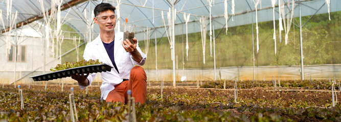 A man in a white lab coat is examining a plant and soil in a greenhouse