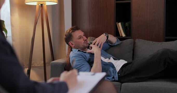 Man lies on couch sharing personal life problems in therapy room. Woman psychologists takes notes in notebook documenting key insights during conversation.