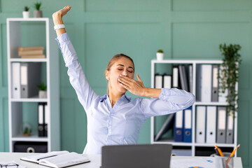 A young lady is sitting at her desk in a modern office. She is yawning and stretching her arms...