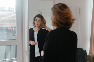 Woman adjusting short wavy hair near mirror, soft room light, relaxed posture, natural expression, simple styling moment near window frame.