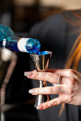A professional bartender's hands pouring vibrant blue liquid from a bottle into a stainless steel jigger. Shallow depth of field with focus on the measuring tool