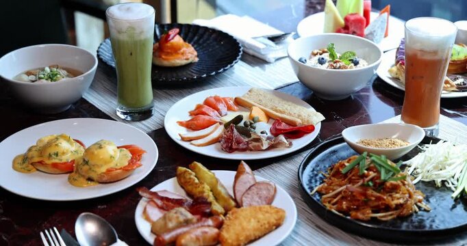 Lavish breakfast buffet spread with diverse dishes: eggs benedict, cold cuts, hot food, drinks. Upscale hotel restaurant setting, bright daylight.