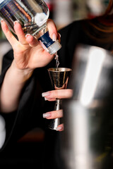 A bartender in a black shirt uses a metal jigger to precisely pour clear alcohol from a glass bottle at a bar counter