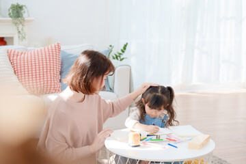 Mother comforts her little daughter drawing or doing homework at a bright, cozy table in the living room, focusing on family love and education.