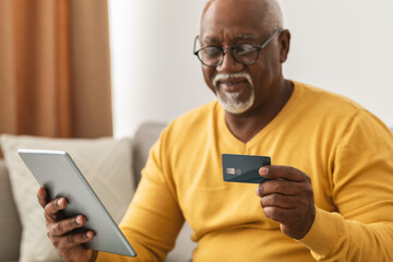 An elderly man sits comfortably on a couch, focused on a tablet in his hands while holding a contactless credit card. Warm lighting creates a cozy atmosphere in the room.