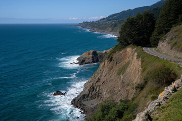 Fototapeta premium The image portrays a scenic coastal cliff with a winding road above, providing a stunning view of crashing ocean waves against rugged cliffs and vibrant greenery along the coast.