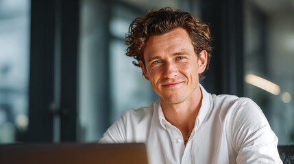 Fototapeta premium Close-up portrait of a smiling man with curly hair. He's wearing a white shirt and is working on his laptop. Modern workplace, focused, confident, approachable, and engaging.
