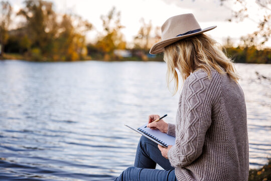 Woman sitting by lake, writing into notebook or diary while enjoying tranquil moment. Personal growth intentions during calm journaling outdoors
