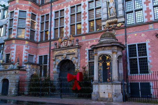 The Great Armoury is a 17th-century former arsenal, Mannerist architecture in Gdansk, Poland. A large red bow hangs on the entrance. The scene is captured during the evening with soft light.