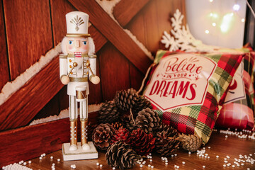 Classic Christmas Still Life Featuring a White and Gold Nutcracker Figure Standing Beside a Pile of Pinecones and a Plaid Pillow with the Text 'Believe in your Dreams' on a Wooden Background