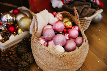 Round Woven Basket Filled with Glittery Pink and Gold Christmas Ornaments and White Fluffy Material, with Pinecones and Other Decorations Blurred in the Background