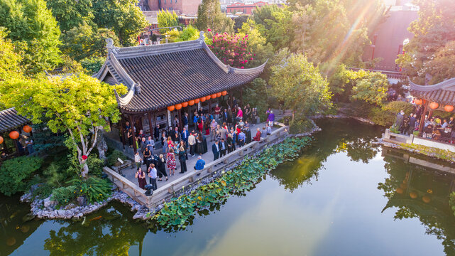 Aerial view of a traditional Chinese pavilion hosting a gathering, its dark tiled roof contrasting with the vibrant greenery and still waters of Lan Su Chinese Garden, Portland, Oregon, United States.