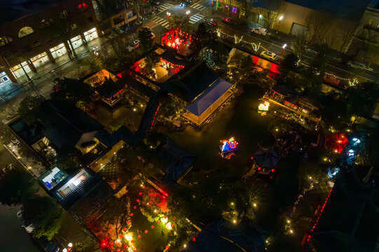Aerial view of the radiant Lan Su Chinese Garden glows with warm lights reflecting off the dark water, a serene contrast to the city's cool tones, Portland, Oregon, United States.