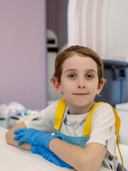 Child seated at worktable in culinary classroom wearing yellow apron and blue disposable gloves. Boy ready to participate in kids' cooking workshop.