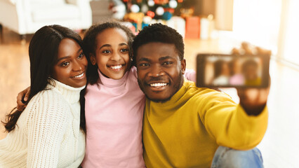 A family sits on the floor, smiling as they take a selfie with a smartphone. They are in a warm indoor space decorated for the holiday season with gifts in the background.