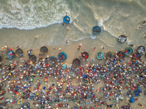 Aerial view of vibrant colors burst as people gather along the sandy shore, their round basket boats dotting the coastline, contrasting with the turquoise waves, Tam Tien, Quang Nam, Vietnam