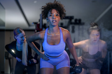Naklejka premium African American and Caucasian women train with dumbbells in a futuristic, blue-lit gym. Intense workout for popular Hyrox and CrossFit health trends, blending fitness with entertainment.
