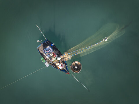 Aerial view of a fishing boat casting its net into the tranquil, dark green waters, its reflection shimmering in the sunlight,Tam Tien, Quang Nam, Vietnam
