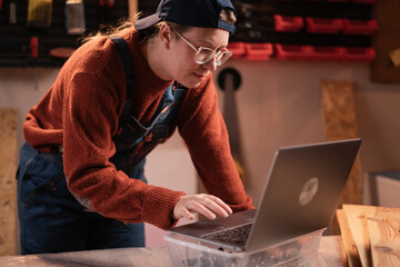 Small business concept. carpenter woman in overalls using laptop computer at woodwork workshop working with laptop computer working in furniture workshop.