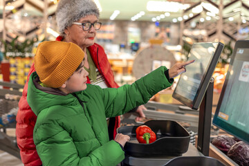 Obraz premium Young boy using self-checkout scale with grandmother supervision at grocery store produce section. modern retail experience, family bonding, digital literacy education and supervised independence 
