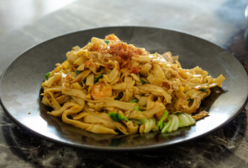 Freshly cooked Indonesian Kwetiau Goreng (Fried Flat Rice Noodles) served hot on a dark plate, topped with fried shallots and shrimp.